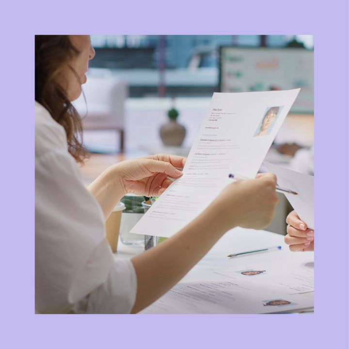 A job candidate's resume being reviewed by a recruiter during an interview. The resume features a profile photo and a combination of skills and experience. The candidate's hands are visible, holding a pen while discussing their qualifications in a professional office setting.