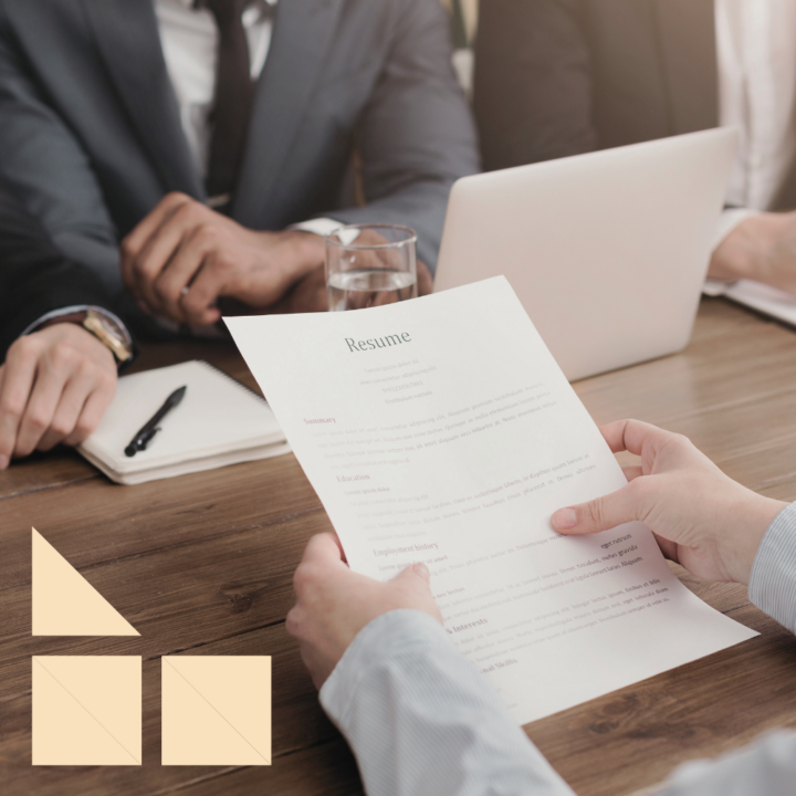 A candidate holding a resume during an interview for an internal promotion. Across the table, interviewers in business attire are engaged in discussion, with a laptop, notebook, and glass of water on the desk, creating a professional setting.
