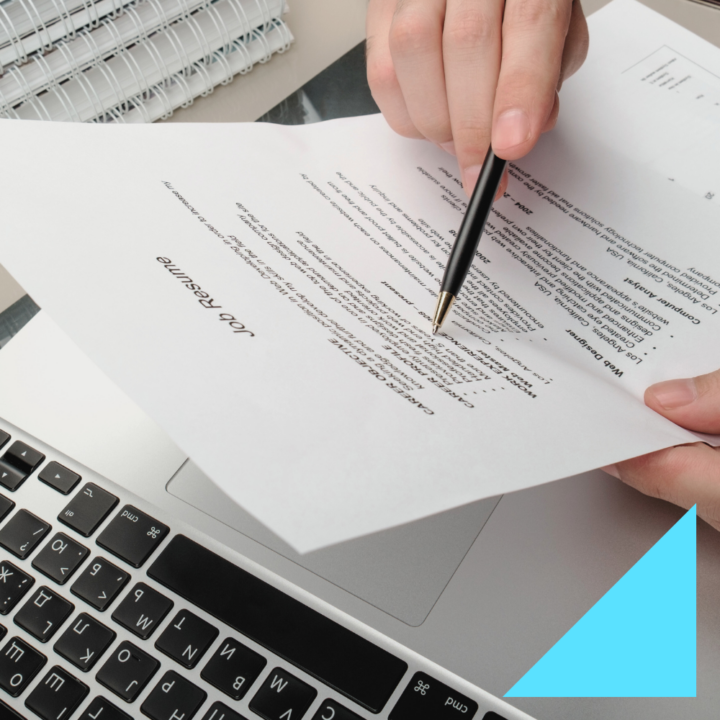 A person reviewing an internship resume with a pen, pointing at key details. The resume is placed over a laptop keyboard, with notebooks and office supplies in the background, creating a professional workspace setting.