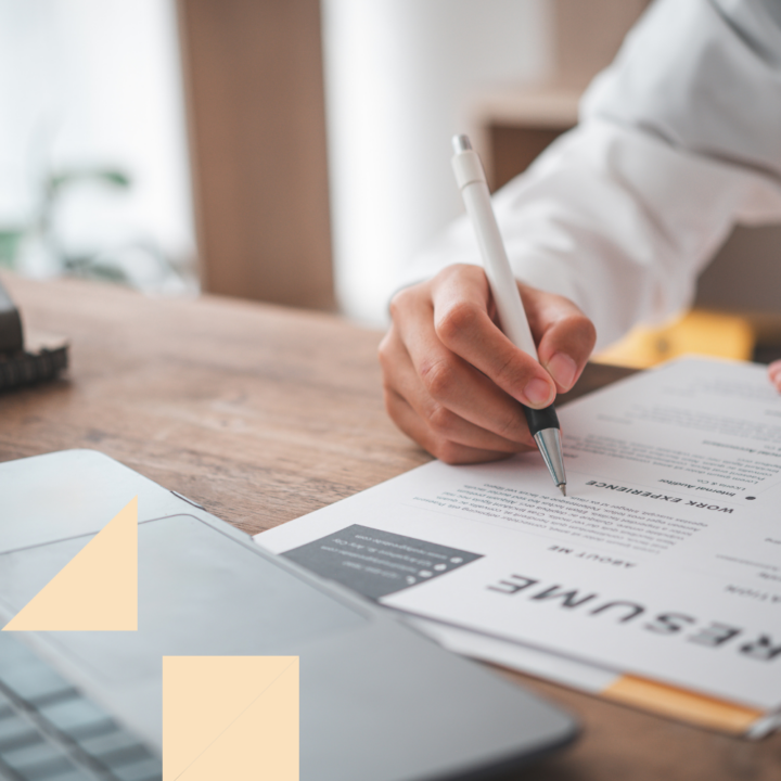 A person in a white shirt reviewing and making notes on a chronological resume. The resume is placed on a wooden desk next to an open laptop, creating a professional and focused workspace.