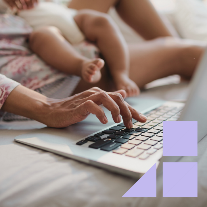A close-up of a mom's hands typing on a laptop while holding a baby. The baby’s legs and feet are visible in the background.