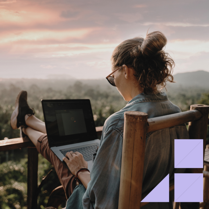 A woman wearing sunglasses and a denim jacket sits on a wooden chair, typing on her laptop while overlooking a scenic tropical landscape at sunset. She has her feet propped up.