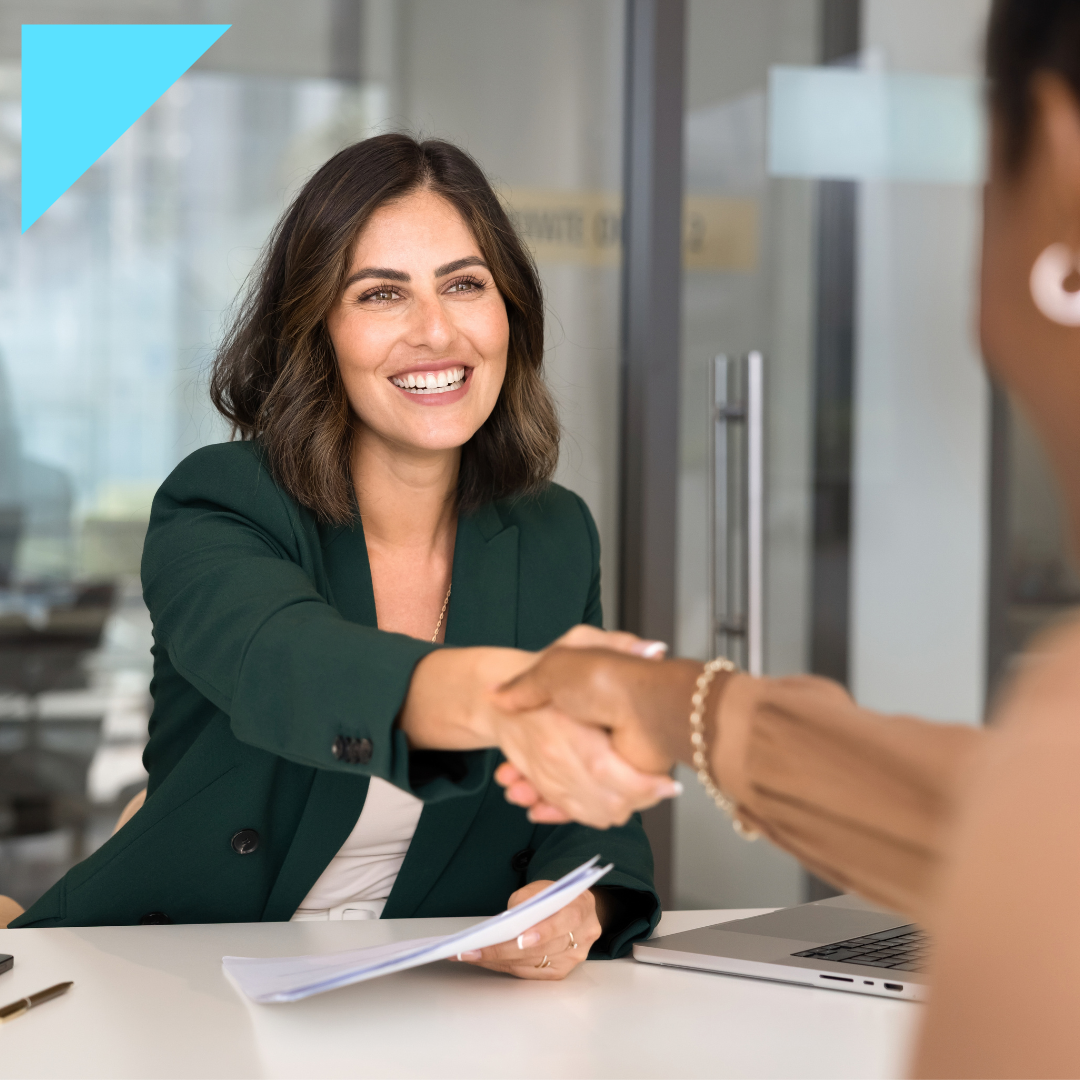 Smiling businesswoman in a green blazer shaking hands with a candidate across a desk during a job interview