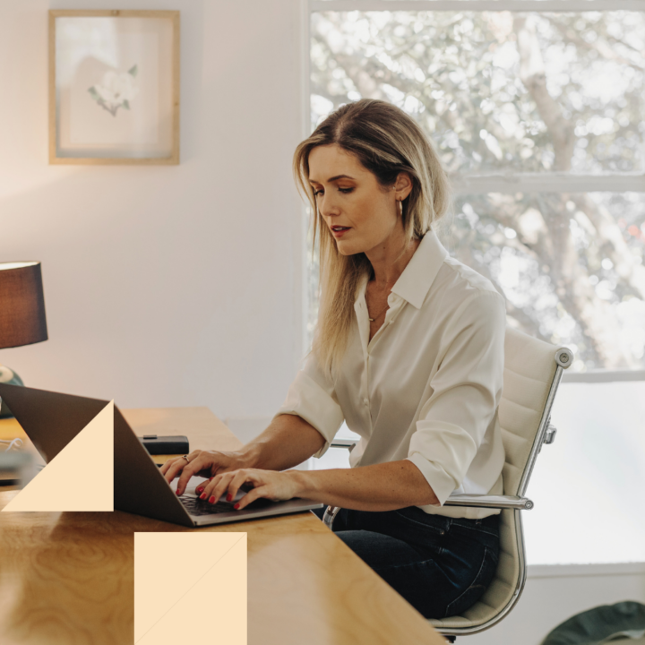 A professional woman with long blonde hair, wearing a white blouse, sits at a wooden desk typing on a laptop. She is focused on her work in a well-lit home office with a large window, framed artwork, and a lamp in the background.