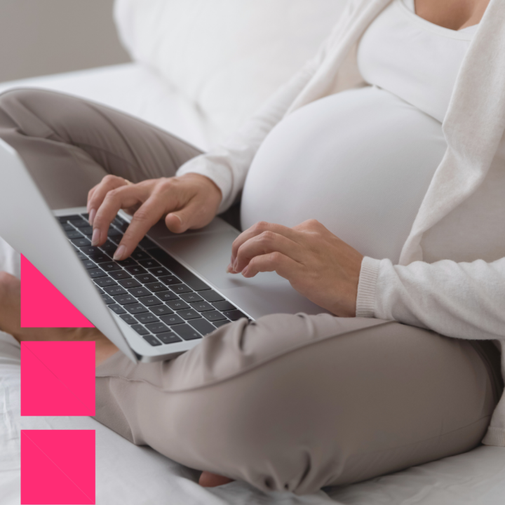 A pregnant woman sitting cross-legged on a bed, typing on a laptop. She is wearing comfortable beige pants and a white top, suggesting she is working or writing from home in a relaxed setting.
