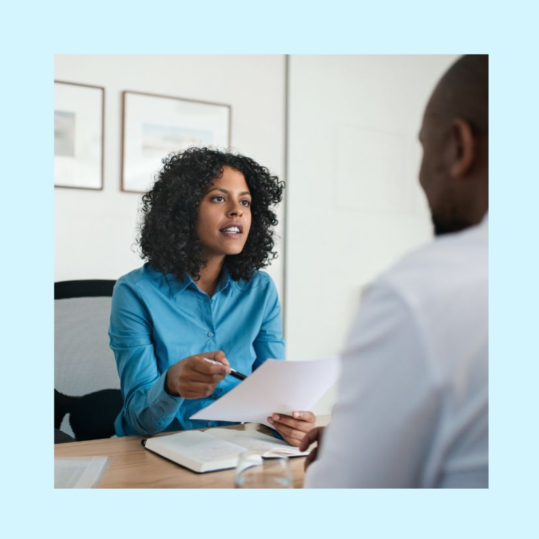 Female hiring manager wearing a blue blouse sitting in her own office across from a candidate holding a piece of paper about to start an interview