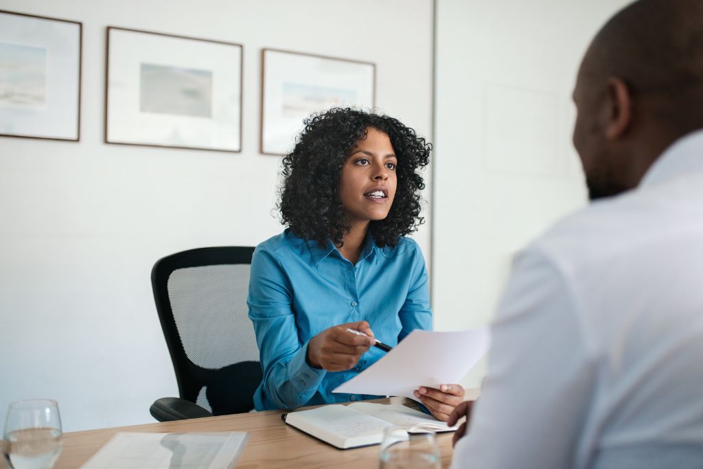 Female hiring manager wearing a blue blouse sitting in her own office across from a candidate holding a piece of paper about to start an interview