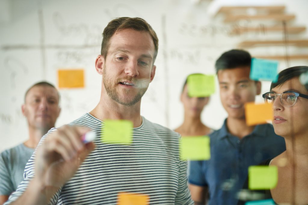Shot of a group of young professionals planning hiring strategy on a glass board writing with pens on sticky notes