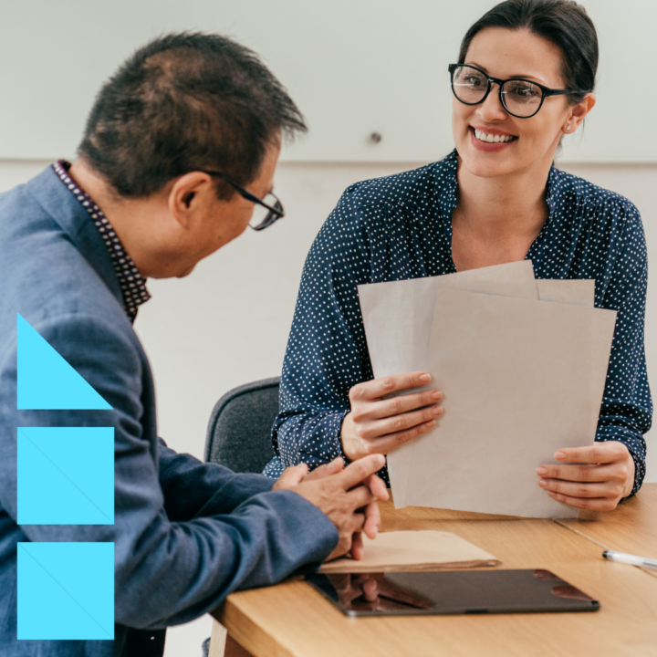 A female executive recruiter wearing glasses and a navy polka dot blouse smiles warmly while reviewing documents with a male candidate seated across from her at a wooden table during a professional interview