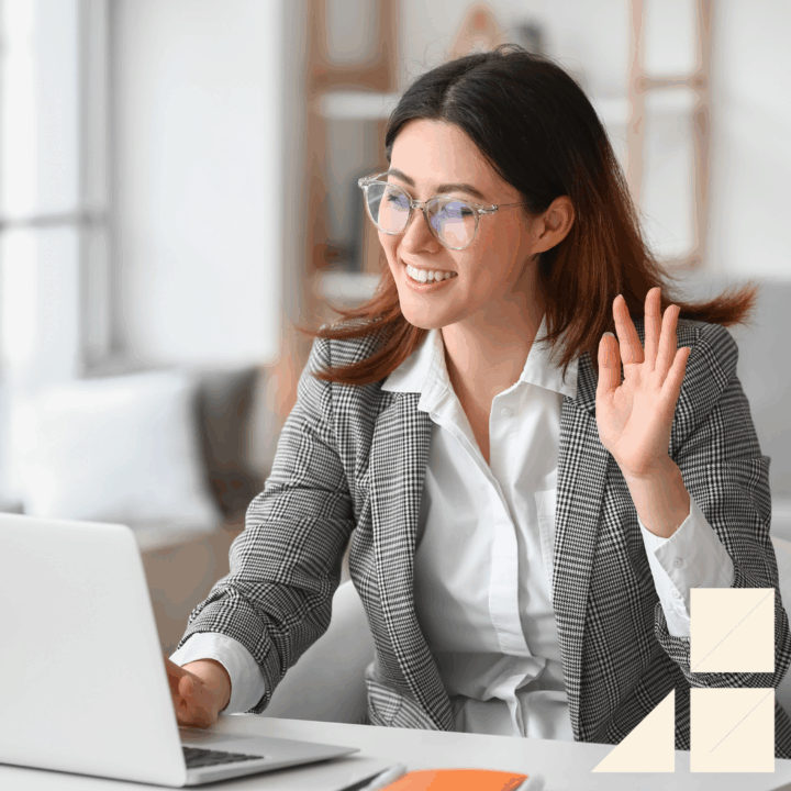 Young female wearing glasses, a white button up dress shirt, and a gray blazer waving at her computer screen getting ready for virtual interview