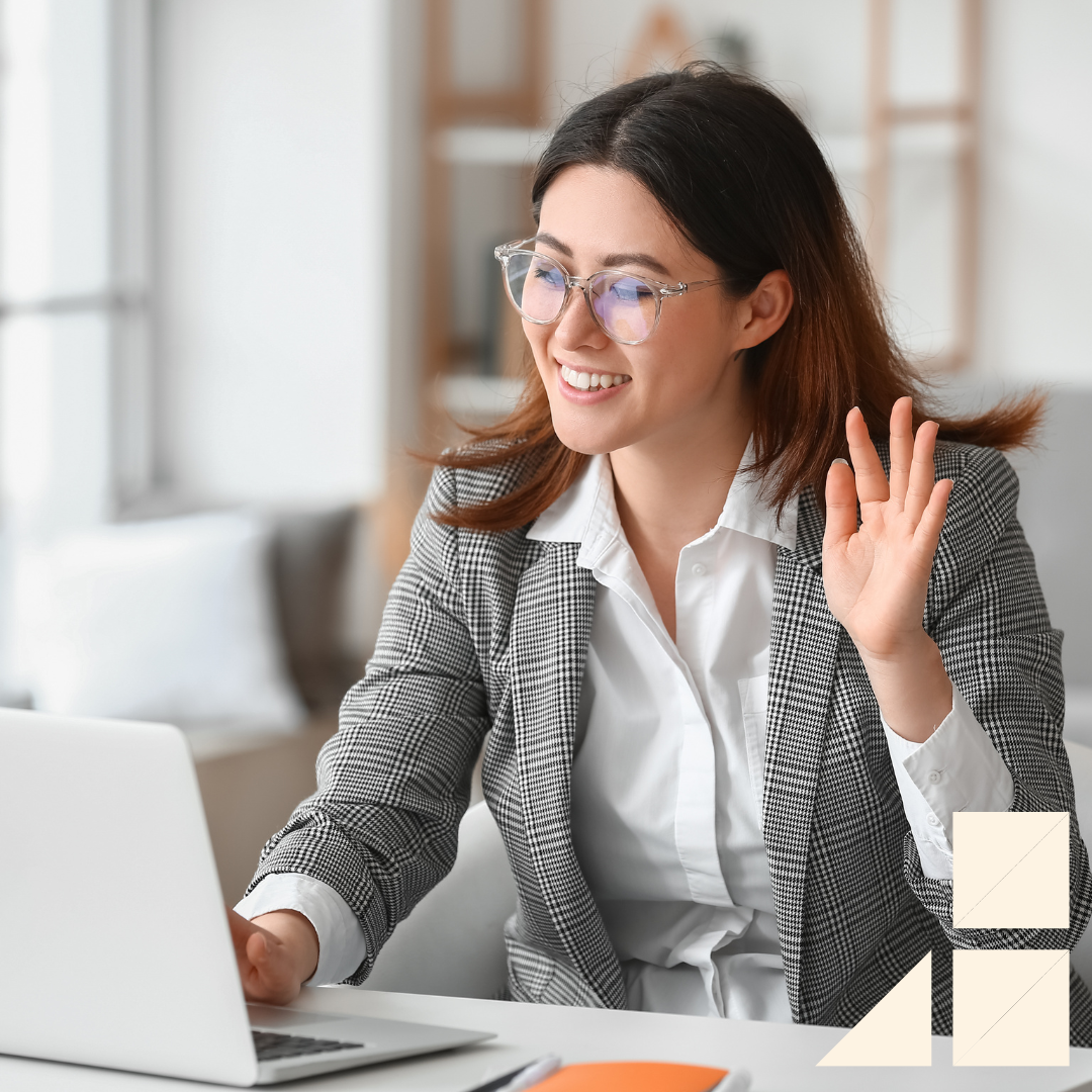 Young female wearing glasses, a white button up dress shirt, and a gray blazer waving at her computer screen getting ready for virtual interview
