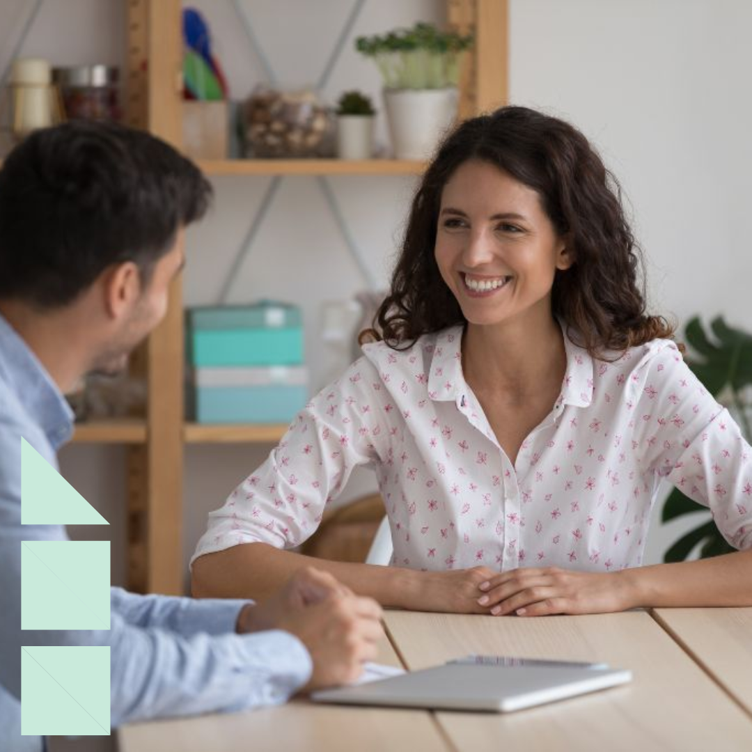 Smiling job candidate in a white patterned blouse speaking with an interviewer across a desk in a bright office, with shelves and plants in the background.