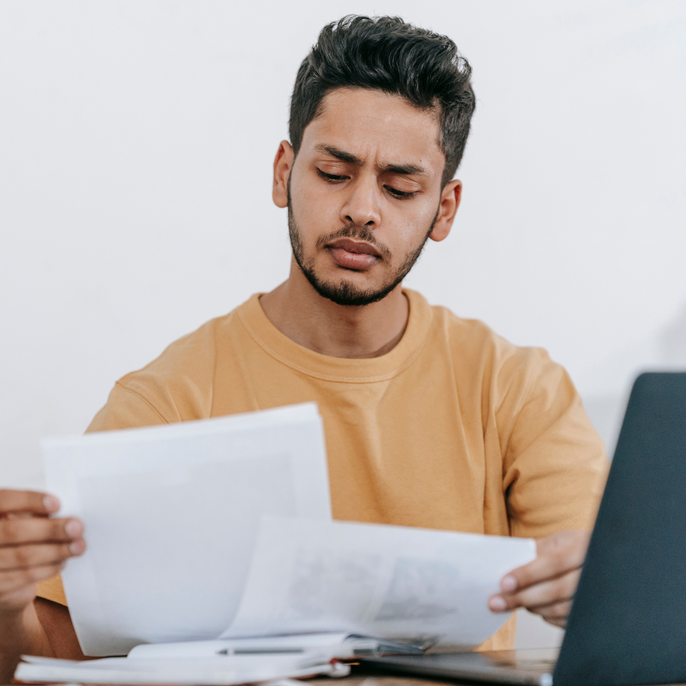 Young male professional with a confused expression sitting behind his laptop looking at print outs of his resume