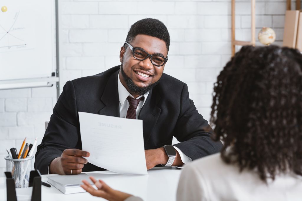 A smiling male interviewer in glasses and a business suit reviews a resume during a job interview, engaging with a female candidate whose back is to the camera in an office setting.