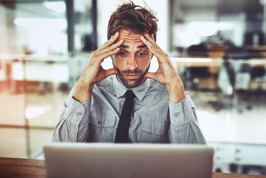 Male professional in a button up and tie in a busy office sitting at his desk in front of his laptop with his hands on his temples and looking confused