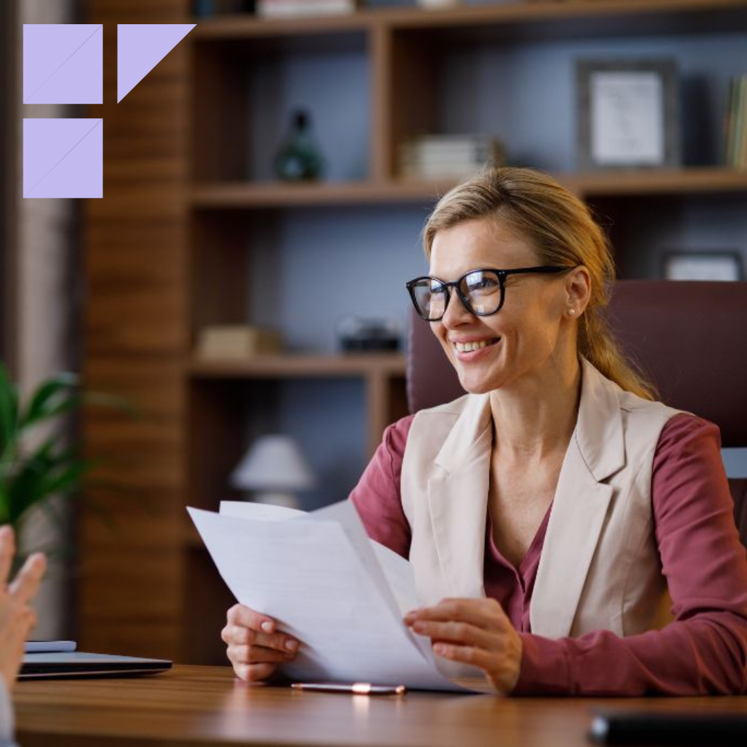 Female hiring manager for a small business smiling wearing black glasses and a beige blazer is sitting in a modern office across from job candidate during an interview
