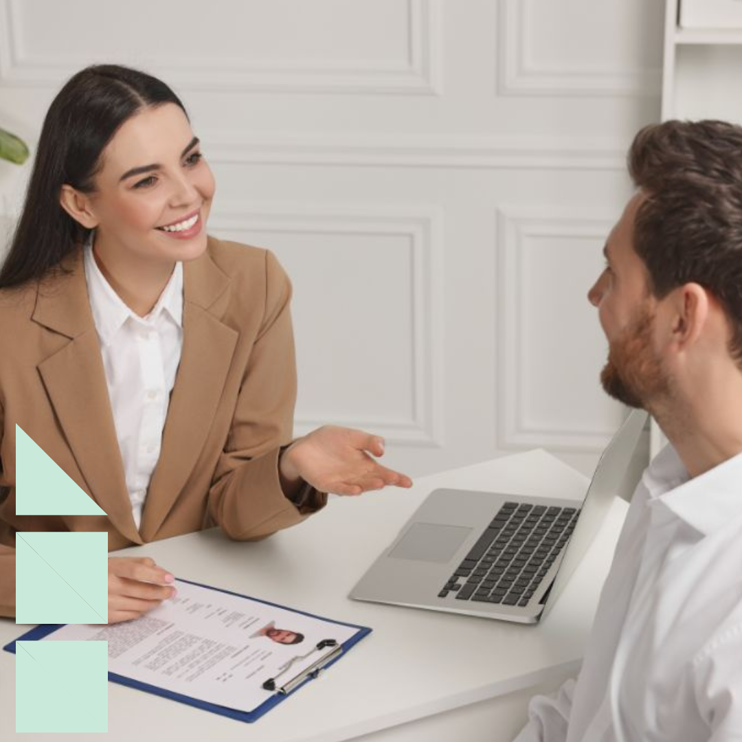 A professional job interview setting featuring a talent acquisition woman in a beige blazer conducting an interview with a male candidate. The woman is smiling and engaged, holding a clipboard with a resume visible on top. The man, seated across from her, appears attentive and is dressed in a white shirt. A laptop is open in front of the woman on a white desk, which also has a pen and notebook. The background includes white paneled walls and office shelves, creating a formal and organized atmosphere.