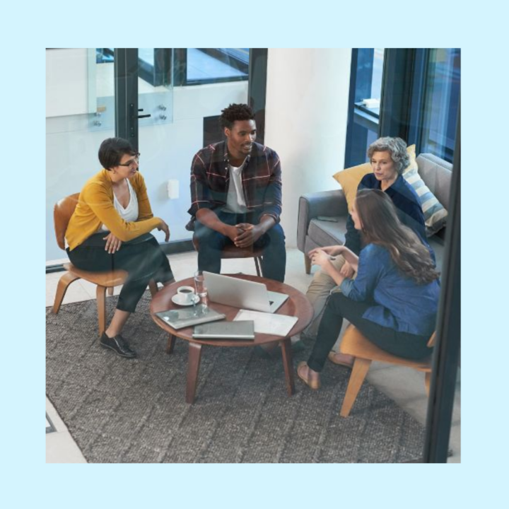A group of four people is seated in a modern office lounge, engaged in a casual meeting about partnering with staffing agency. Two women and two men sit around a small round table with laptops and coffee cups. One woman in a mustard-yellow sweater leans forward, while another gestures as she speaks. The others listen attentively, creating a collaborative and relaxed atmosphere with glass walls and cozy seating.