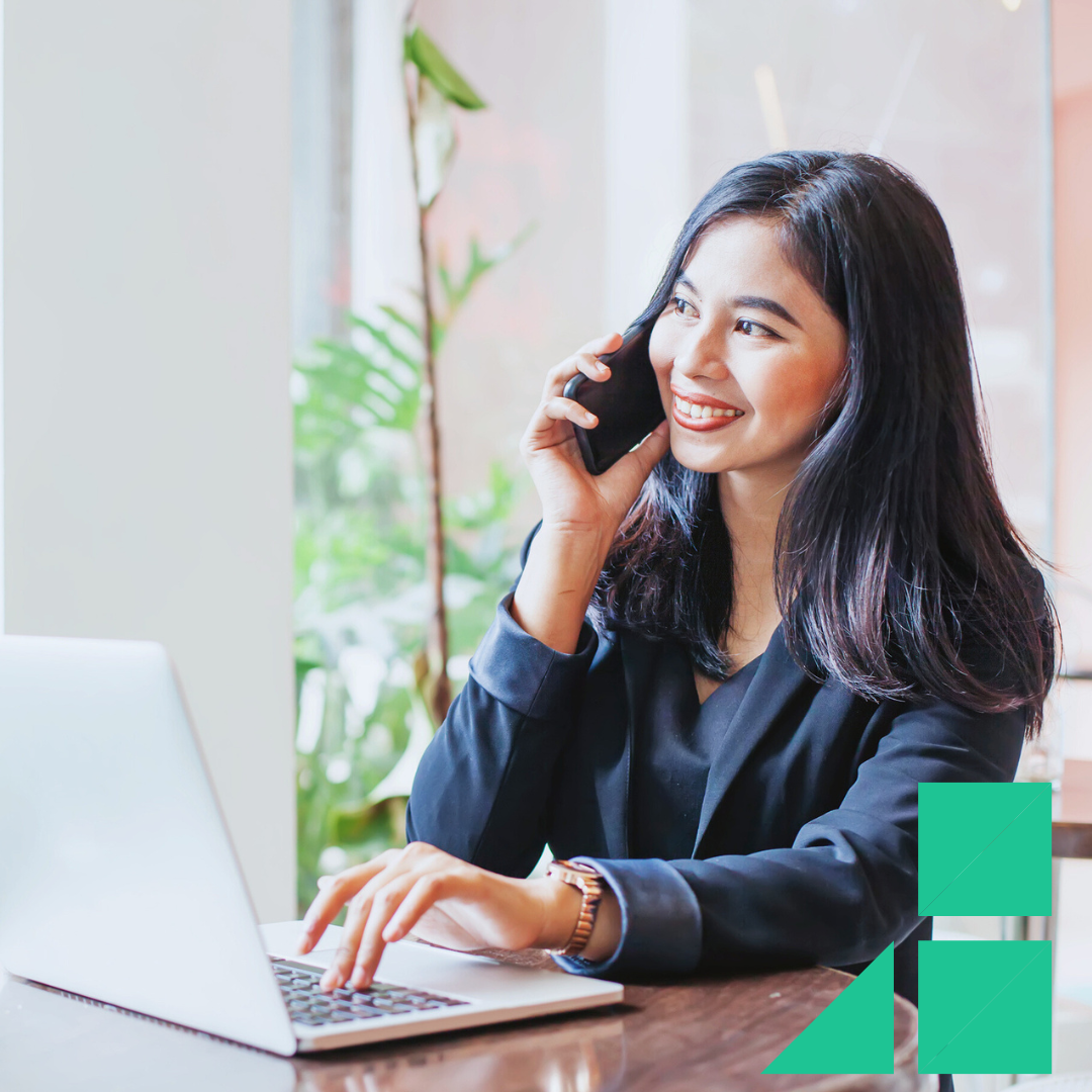 A young professional woman smiling while participating in a phone interview, sitting at a table with a laptop in front of her. The setting is bright and modern, with plants and large windows in the background.