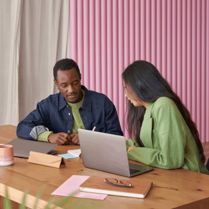 An intern and a manager collaborating at a wooden desk. The intern attentively listens to the manager, who is explaining something on a laptop screen. The background features pink vertical blinds.