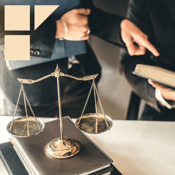 Close-up of brass scales of justice placed on top of stacked legal books, with two professionals in business attire discussing a legal document in the background; a beige geometric logo is displayed in the upper left corner.