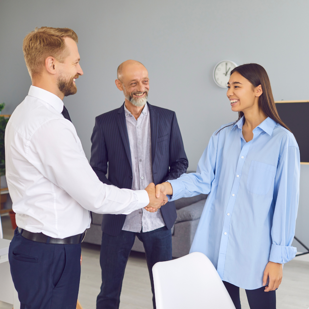 Young professional woman in a light blue shirt shaking hands with a man in a white dress shirt during a business meeting, while another man in a striped blazer smiles and observes in the background, indicating a positive introduction or job interview setting.