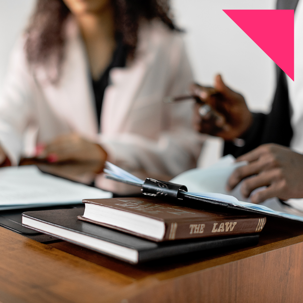 Two people in class at a desk with law-related books, one titled 'The Law,' and papers. One person is holding a pen, and there are pink highlights or marks drawn on the image, emphasizing the focus on law and study materials.