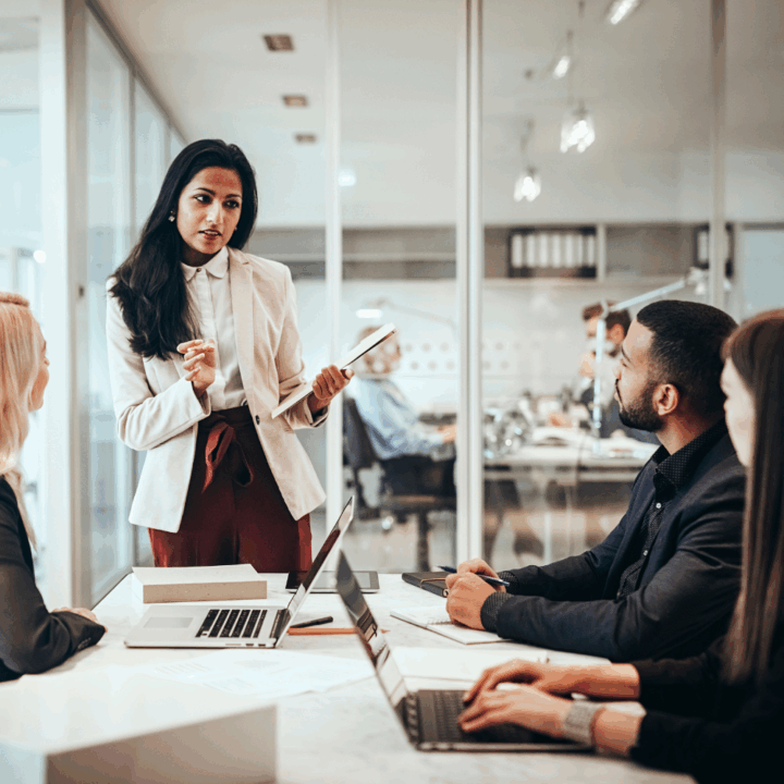 Professional woman confidently talking to colleagues in a modern office meeting room, navigating a conversation with difficult coworkers, as laptops and documents sit on the table