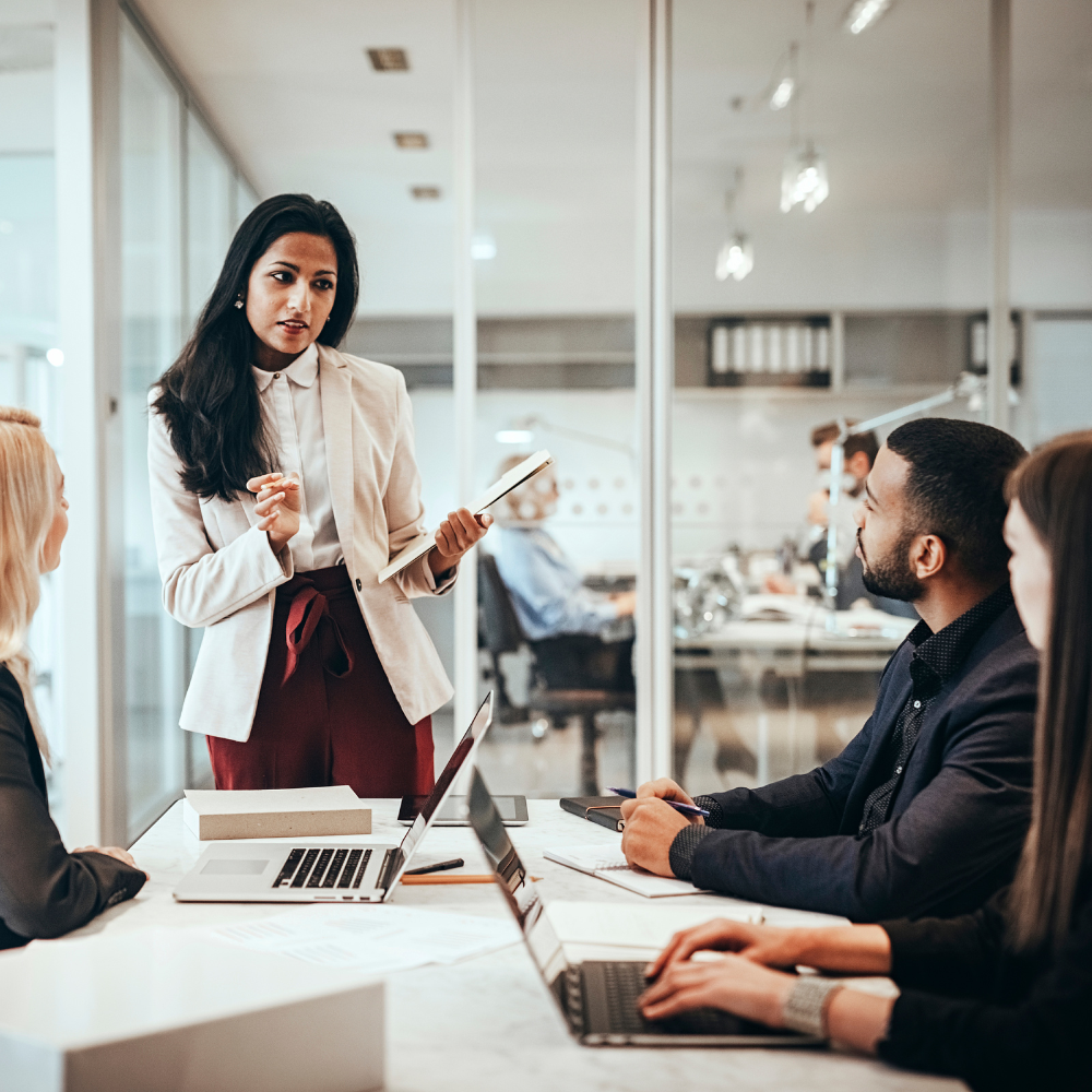 Professional woman confidently talking to colleagues in a modern office meeting room, navigating a conversation with difficult coworkers, as laptops and documents sit on the table