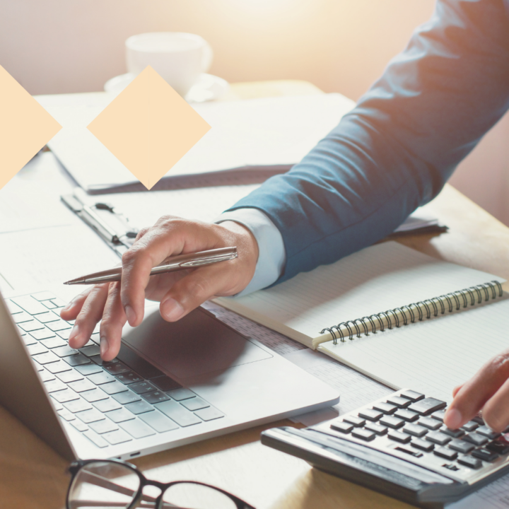 A person in a business suit working at a desk, typing on a laptop with one hand while using a calculator with the other. A pen is held in the typing hand, and a spiral notebook lies open nearby