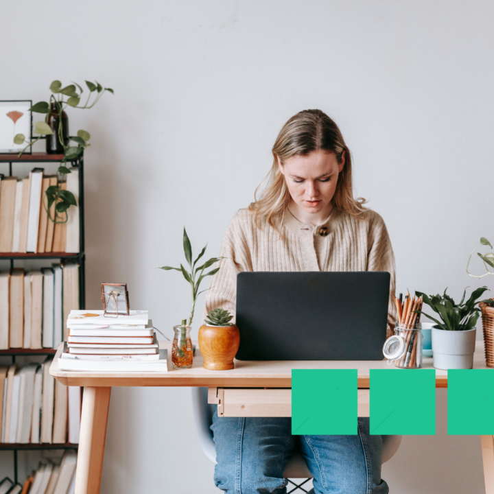 Young woman working on a laptop at a desk with books and plants, focused on completing an online marketing course.
