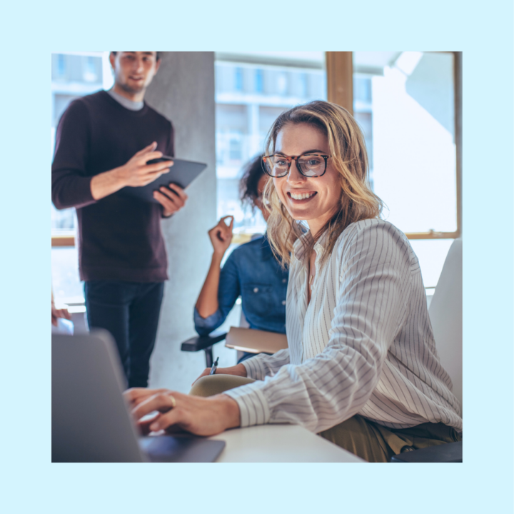Smiling woman with glasses working on a laptop in an office, with colleagues interacting in the background.