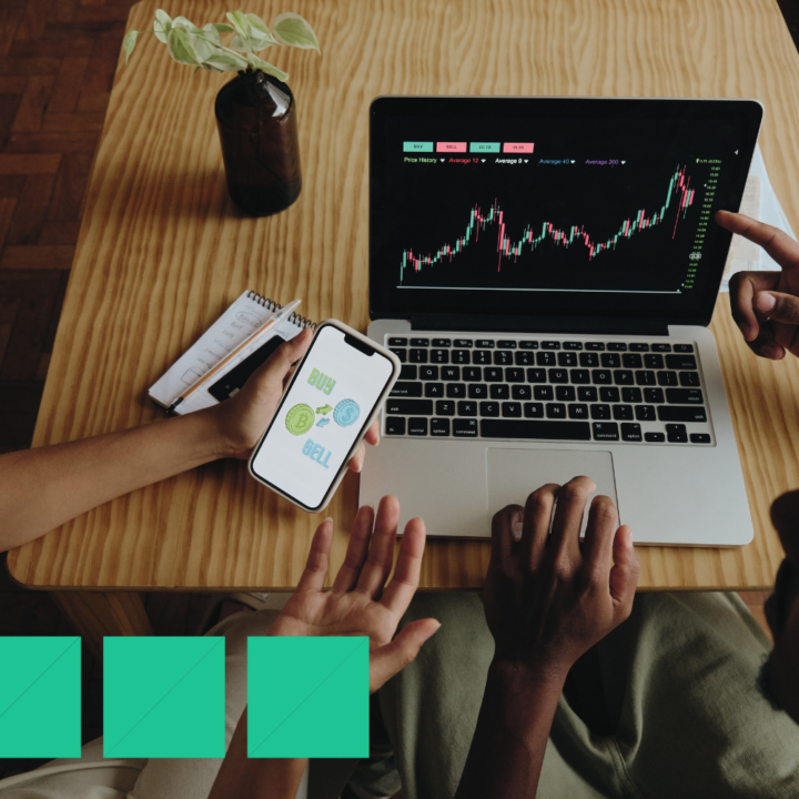 Employees analyzing cryptocurrency market data on a laptop screen while discussing buy and sell options displayed on a smartphone, seated at a wooden desk with a notepad and plant.