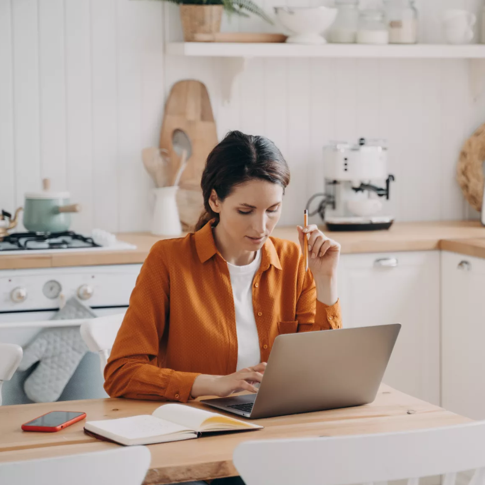 Female freelancer on her laptop next to a notepad at a table in her kitchen