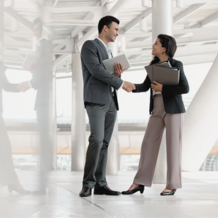 Female business woman standing holding a binder introducing herself to business man. Both in professional attire shaking hands in the office.