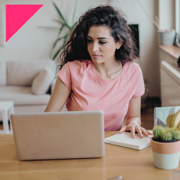 A young woman with curly hair wearing a pink t-shirt is sitting at a wooden table, passively job searching on a laptop with a notebook beside her. She appears focused and engaged, with a comfortable home setting in the background.