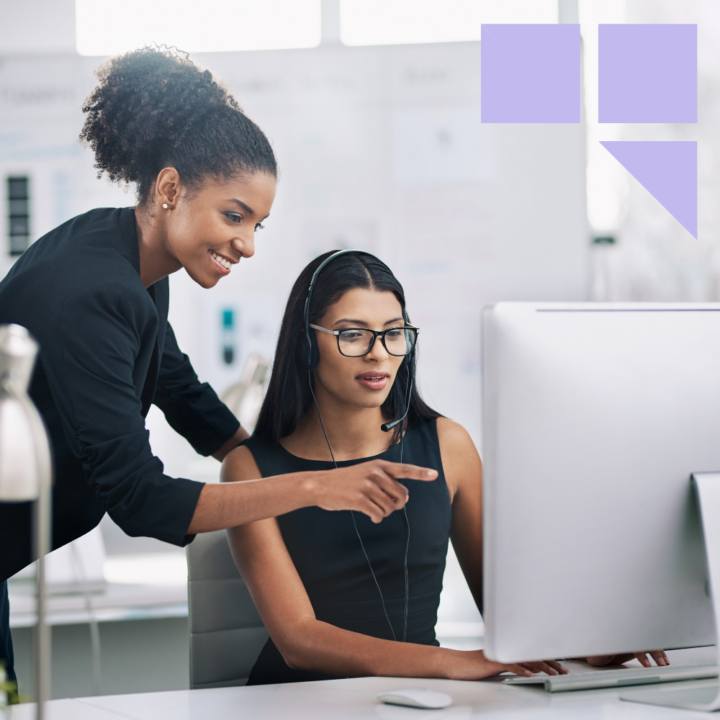 A new manager offering hands-on support to an employee wearing a headset, as they work together at a computer in a modern, well-lit office setting.