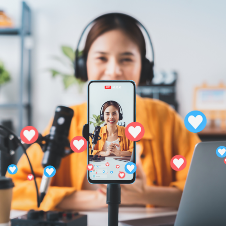 A woman wearing headphones and a yellow jacket conducts a live social media broadcast, speaking into a microphone while being recorded on a smartphone. Floating heart icons indicate audience engagement.