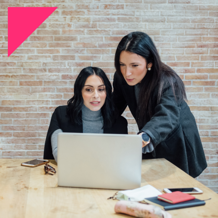 Two women with dark hair are working together at a desk, looking intently at a laptop screen. One woman is sitting while the other is standing, pointing at the screen.