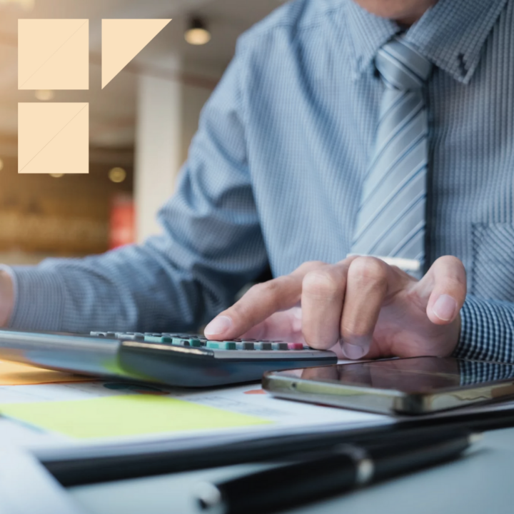 An accounting male professional wearing a blue-striped shirt and tie, working on a calculator with documents and a smartphone on the desk.