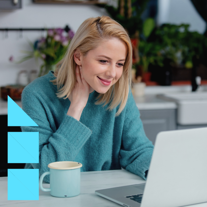 A woman with blonde hair wearing a teal sweater smiles while working on her laptop at a kitchen table, with a coffee mug beside her and plants in the background.