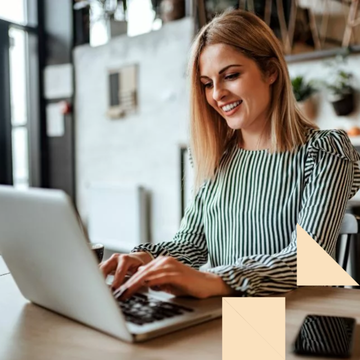 Smiling woman typing a cover letter and letter of interest on laptop indoors sitting at a table next to her phone.