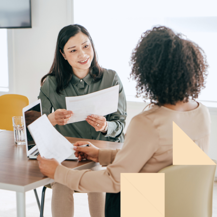 Two professional women engaged in a job interview or business discussion in a modern office setting. One woman, wearing a green blouse, is smiling while holding a document. The other woman, dressed in a beige top, is taking notes on a paper.