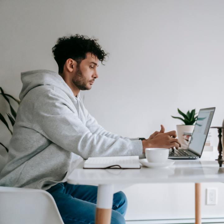 A young man in a gray hoodie sits at a white table, focused on his laptop. A notebook and coffee cup are nearby, suggesting he is composing an important message, possibly declining a job offer.