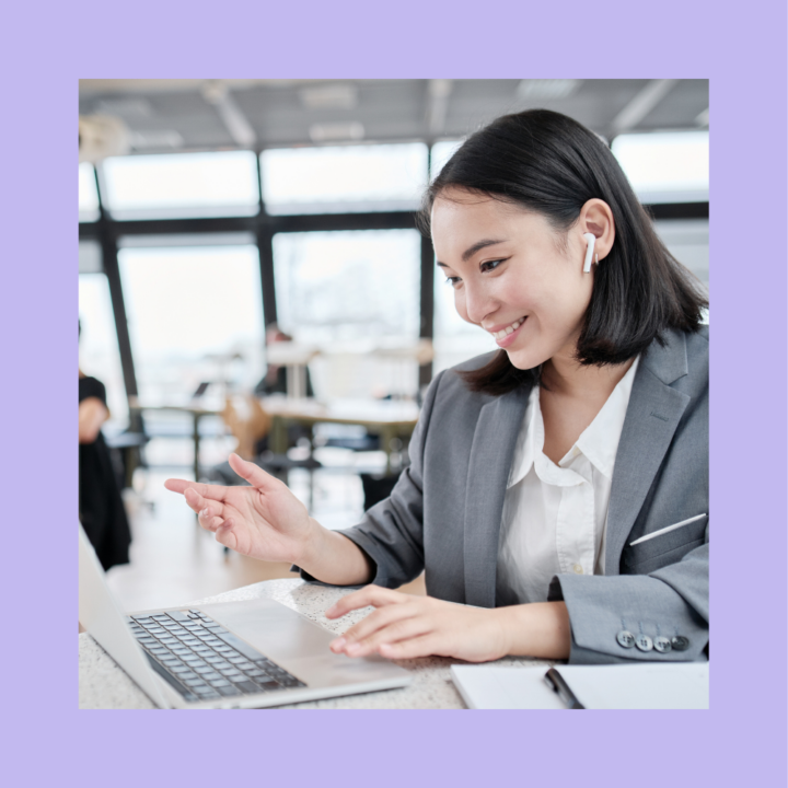 A professional recruiter woman in a gray blazer and white blouse engages in a virtual interview, smiling and gesturing towards her laptop screen. She is wearing wireless earbuds and sitting in a modern office space with large windows and blurred colleagues in the background.
