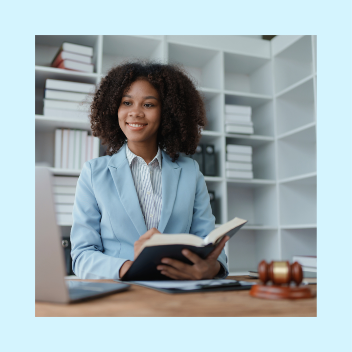 A young woman in a light blue blazer sits at a desk in a law office, holding an open book and smiling. Behind her are shelves filled with books and files. A laptop and a judge’s gavel are on the desk, suggesting a legal or professional internship setting.