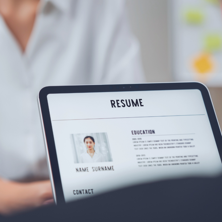 A close-up of a tablet displaying a digital resume, with a professional headshot and education details visible. In the blurred background, a person in a white shirt appears to be reviewing the resume, possibly during an interview or hiring process.