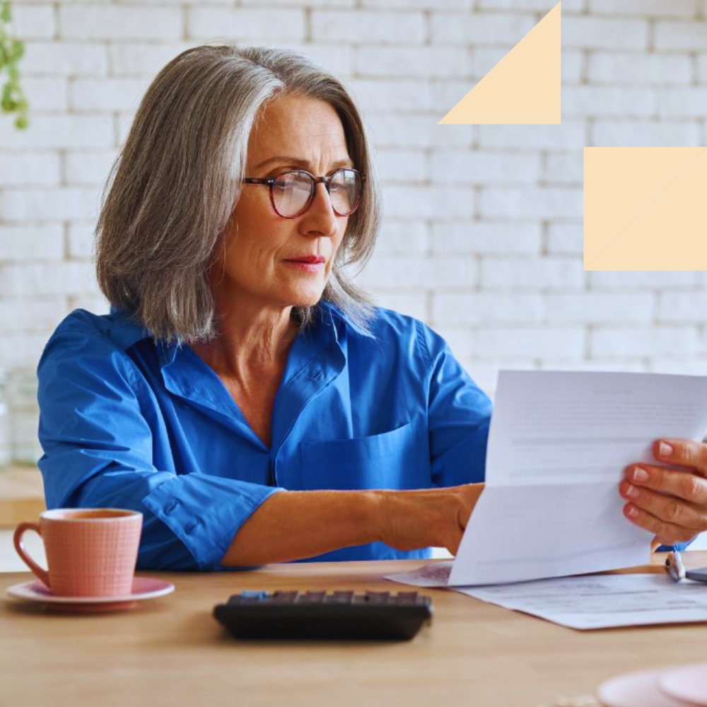 An older woman with gray hair and glasses, wearing a blue blouse, sits at a wooden table reviewing a document. A calculator, paperwork, and a cup of coffee are placed in front of her, suggesting she is analyzing financial or retirement-related information.