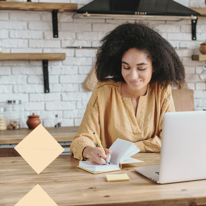 A woman with curly hair wearing a mustard-colored blouse sits at a wooden kitchen table, writing in a notebook with a pencil while using a laptop. She appears focused and engaged, with a slight smile, in a cozy, modern kitchen setting.