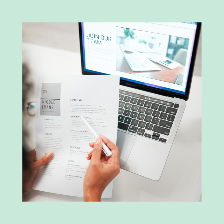 A person holding a printed mid-career resume while reviewing it with a pen, sitting at a desk with an open laptop displaying a "Join Our Team" recruitment webpage. The resume includes sections on experience, education, and awards.
